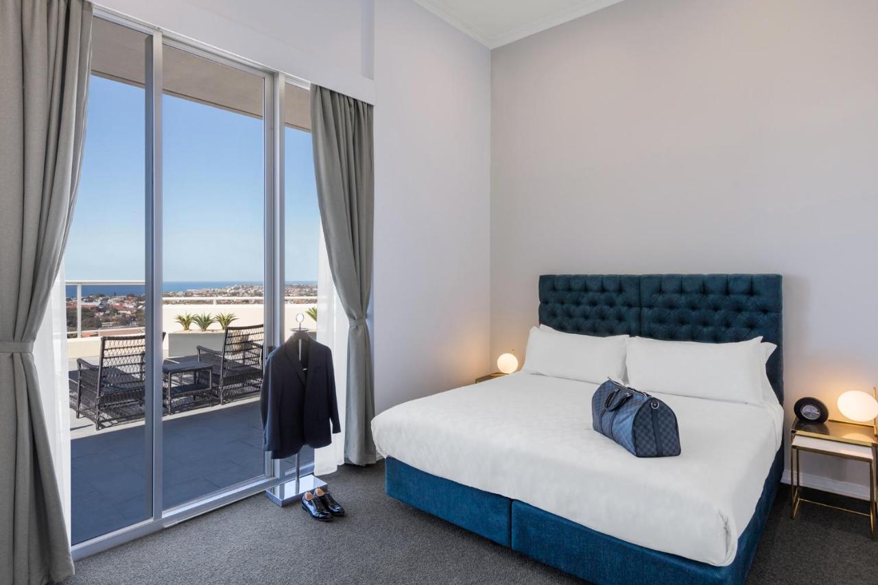 فندق أجنحة ميريتون بوندي جنكشن سيدني المظهر الخارجي الصورة The photo shows a stylish bedroom featuring a large bed with a tufted blue headboard. There's a neatly organized outfit consisting of a suit hanging on a stand, along with shoes placed on the floor. The room has large windows that provide a view of t
