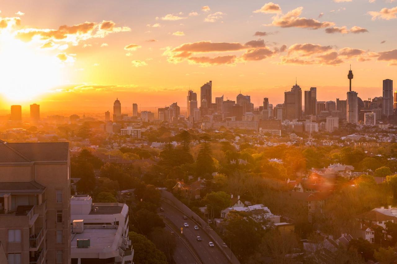 فندق أجنحة ميريتون بوندي جنكشن سيدني المظهر الخارجي الصورة The photo shows a panoramic view of a city skyline at sunset. The sun is low on the horizon, casting a warm golden light across the buildings. Tall skyscrapers are silhouetted against the colorful sky, which features hues of orange, pink, and yellow,