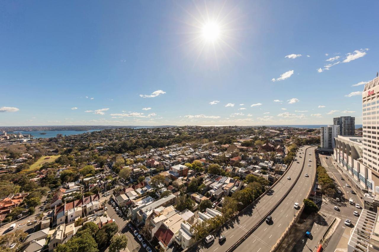 فندق أجنحة ميريتون بوندي جنكشن سيدني المظهر الخارجي الصورة The photo shows a panoramic view of a city landscape under a clear blue sky. In the foreground, there is a road with vehicles traveling along it, bordered by trees and buildings. In the background, you can see a residential area with various houses, 
