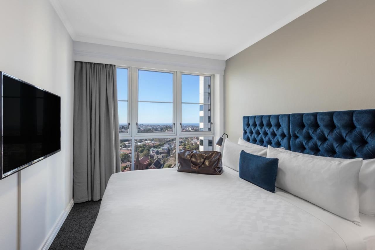 فندق أجنحة ميريتون بوندي جنكشن سيدني المظهر الخارجي الصورة The photo shows a modern hotel room featuring a large bed with a tufted blue headboard. Beside the bed, there is a stylish bag placed on the bed. The room has large windows that provide a view of the outside, allowing natural light to fill the space.