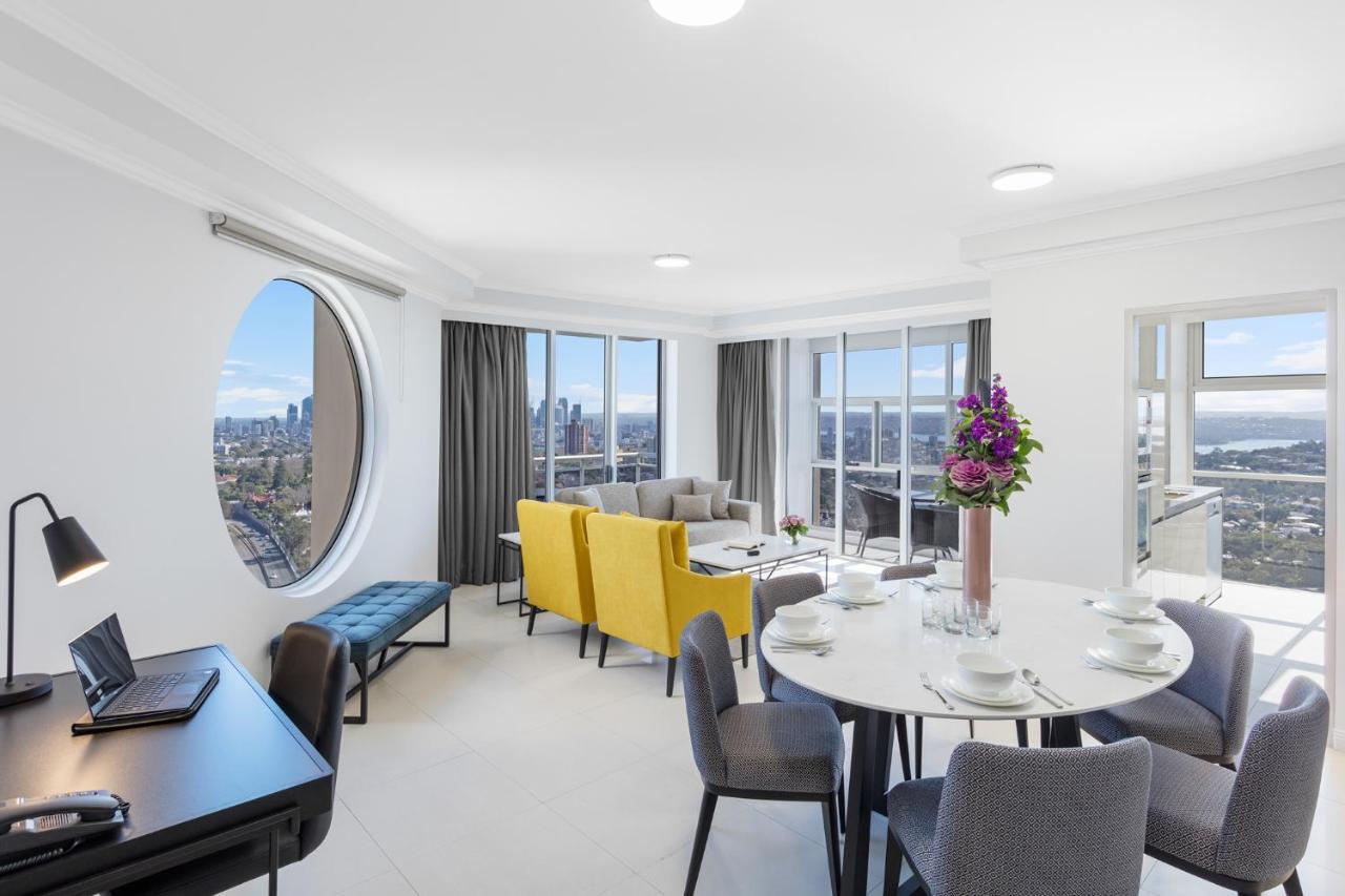 فندق أجنحة ميريتون بوندي جنكشن سيدني المظهر الخارجي الصورة The photo shows a modern and bright interior space, likely a living room or dining area. It features large windows that offer a panoramic view of the city skyline. The furnishings include a round dining table set for several people, with plates and g