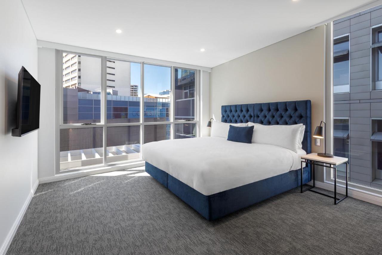 فندق أجنحة ميريتون بوندي جنكشن سيدني المظهر الخارجي الصورة The image shows a modern hotel room with a large, comfortable bed featuring a blue upholstered headboard. The room has large windows that allow natural light to enter, providing a good view of the surrounding buildings. There is a small bedside table
