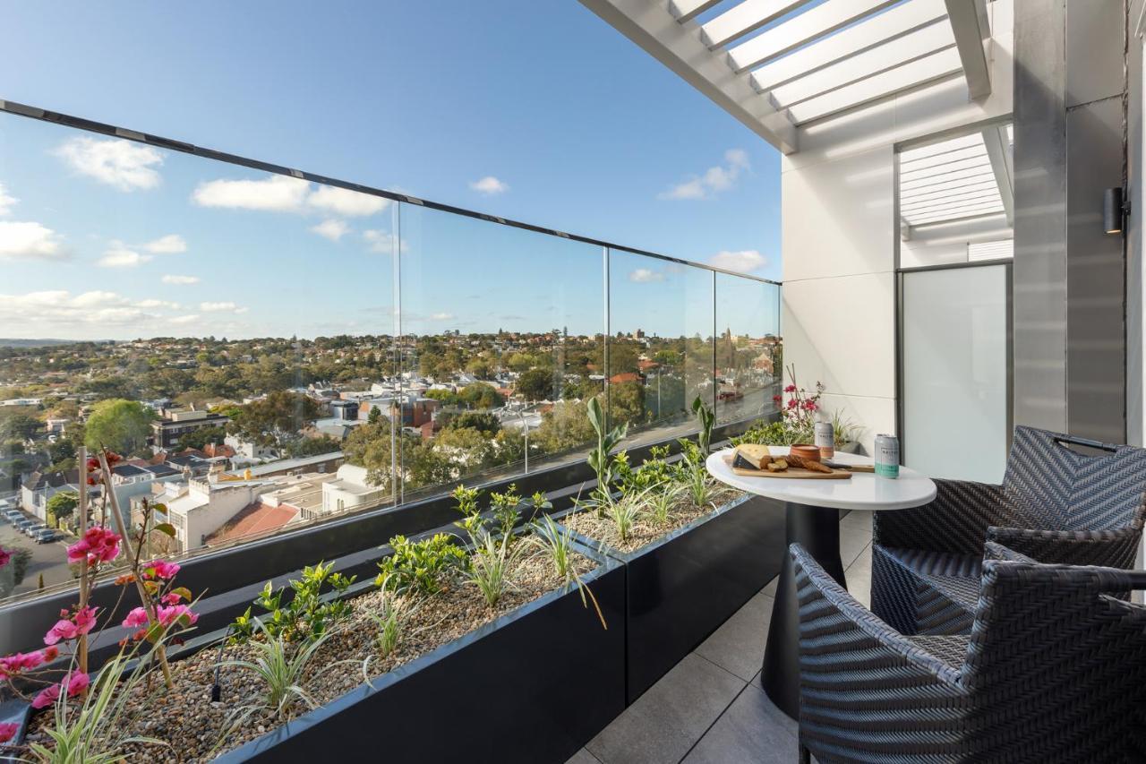 فندق أجنحة ميريتون بوندي جنكشن سيدني المظهر الخارجي الصورة The photo shows a modern balcony with a glass railing, enhancing the view of the surrounding area, which includes various rooftops and greenery in the distance. There is a small round table set with a decorative floral arrangement and some items, lik