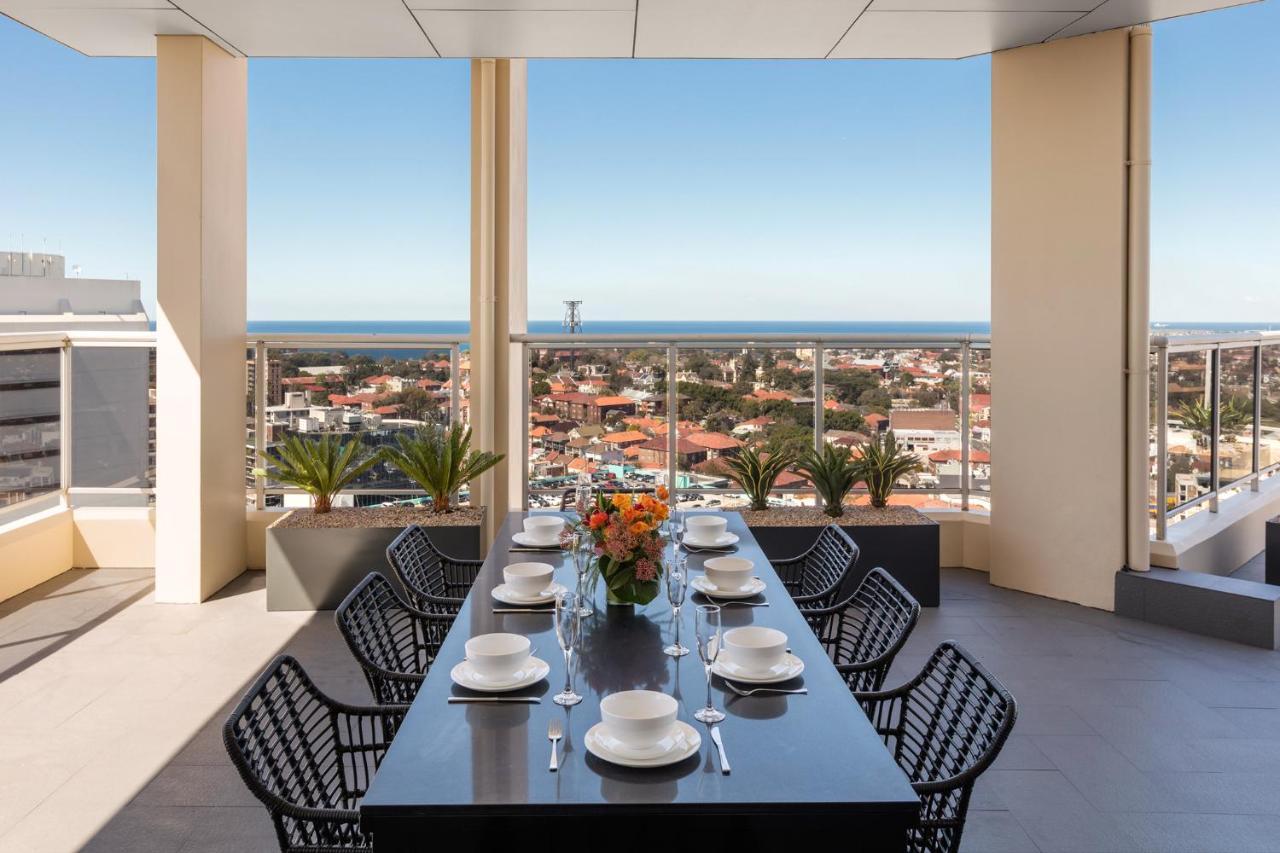 فندق أجنحة ميريتون بوندي جنكشن سيدني المظهر الخارجي الصورة The photo shows a modern outdoor dining area situated on a balcony or terrace with a stunning view. There is a long rectangular table set with plates, glasses, and cutlery, ready for a meal. The table is surrounded by sleek, black wicker chairs. In t