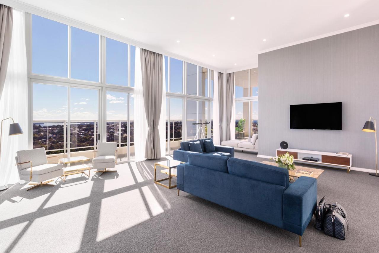 فندق أجنحة ميريتون بوندي جنكشن سيدني المظهر الخارجي الصورة The photo shows a bright and modern living room with large floor-to-ceiling windows that provide a view of the outdoors. The space features a blue sofa in the center, complemented by light-colored armchairs nearby. There are light curtains hanging by