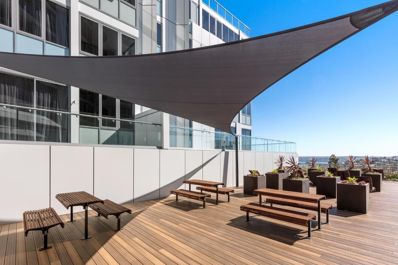 فندق أجنحة ميريتون بوندي جنكشن سيدني المظهر الخارجي الصورة The photo shows a modern outdoor terrace area on a building. It features several wooden benches arranged on a wooden deck. There are large planters with greenery, adding some decorative elements. A large triangular gray shade sail is positioned above