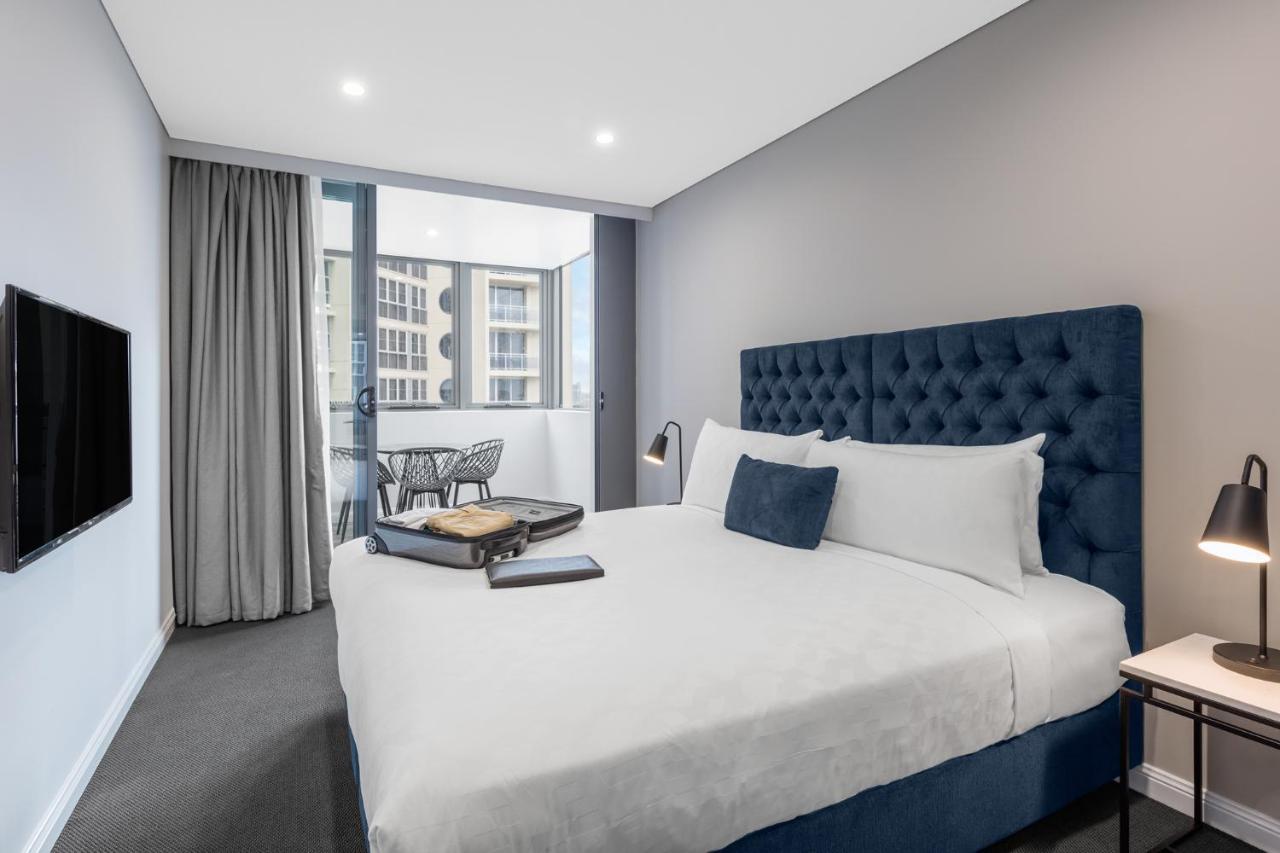 فندق أجنحة ميريتون بوندي جنكشن سيدني المظهر الخارجي الصورة The photo shows a modern hotel room featuring a large bed with a tufted blue headboard. There are minimalistic bedside tables with lamps on either side of the bed. A flat-screen TV is mounted on the wall across from the bed. To one side, there are fl