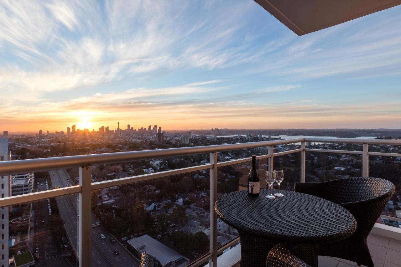 فندق أجنحة ميريتون بوندي جنكشن سيدني المظهر الخارجي الصورة The image shows a scenic view from a balcony or terrace overlooking a cityscape during sunset. In the foreground, there is a small round table with two glasses and a bottle of wine, suggesting a romantic or relaxing setting. The sky is filled with vi