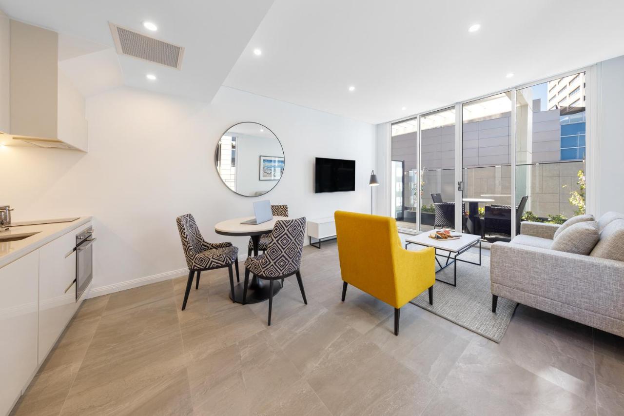 فندق أجنحة ميريتون بوندي جنكشن سيدني المظهر الخارجي الصورة The photo shows a modern and stylish living space. On one side, there is a kitchen area with sleek cabinetry, and next to it, a round dining table surrounded by three patterned chairs. In the center of the room, there is a gray sofa accompanied by a 