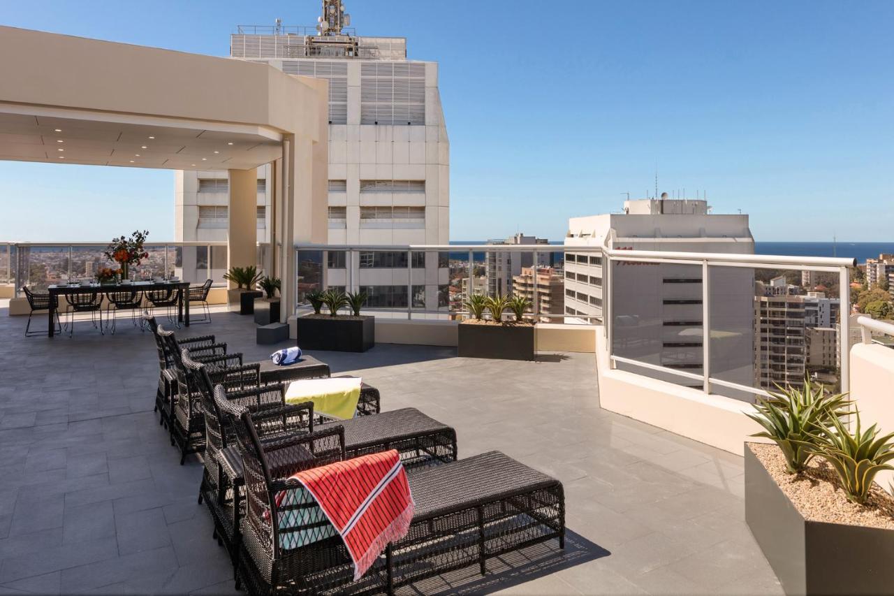 فندق أجنحة ميريتون بوندي جنكشن سيدني المظهر الخارجي الصورة The photo shows a rooftop terrace with a modern design. There are several outdoor seating arrangements featuring black wicker lounge chairs. Some of the chairs have colorful blankets draped over them. The terrace is surrounded by a railing and has po