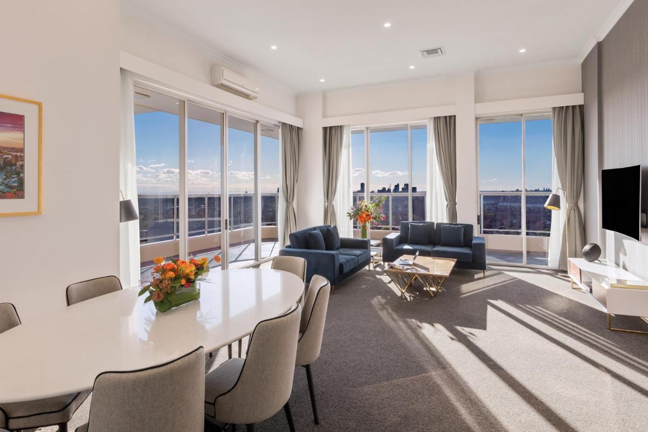 فندق أجنحة ميريتون بوندي جنكشن سيدني المظهر الخارجي الصورة The photo shows a modern and stylish living space with large windows allowing natural light to fill the room. In the foreground, there is a round dining table with several chairs around it, adorned with a floral centerpiece. The seating area features