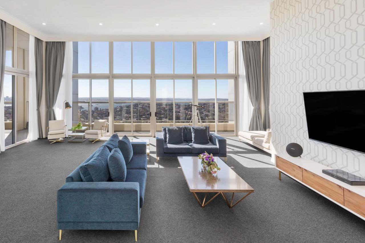 فندق أجنحة ميريتون بوندي جنكشن سيدني المظهر الخارجي الصورة The photo shows a modern and spacious living area. It features large windows that provide a panoramic view of the outside landscape and allow natural light to fill the room. The interior design includes a blue sectional sofa placed in the center, com
