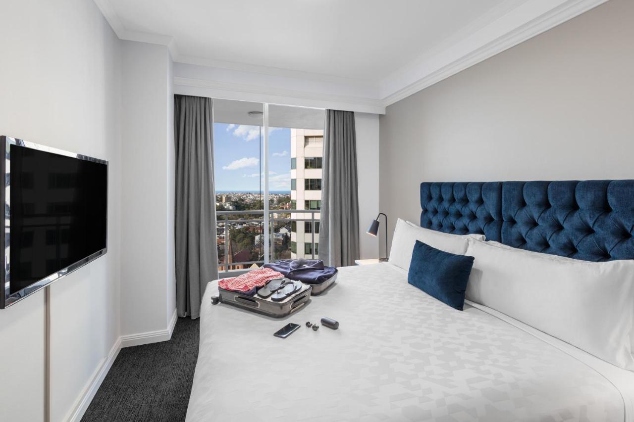 فندق أجنحة ميريتون بوندي جنكشن سيدني المظهر الخارجي الصورة The photo shows a modern hotel room featuring a neatly made bed with a blue tufted headboard and a white duvet. There's a small decorative pillow on the bed, and a piece of luggage is placed at the foot with clothes partially unpacked. A television i