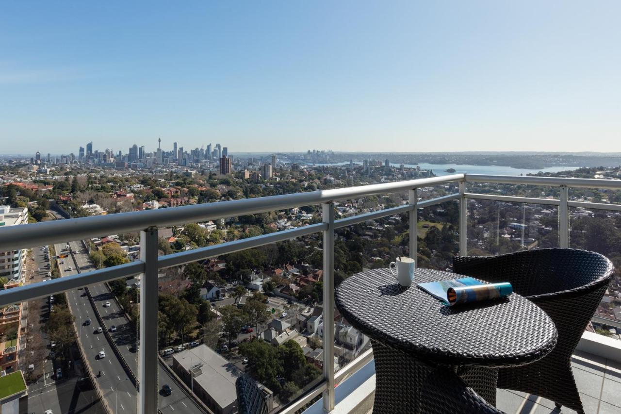 فندق أجنحة ميريتون بوندي جنكشن سيدني المظهر الخارجي الصورة The photo shows a balcony with a view overlooking a cityscape and a body of water in the distance. There is a round table with a woven design and two chairs, one of which has a mug on it. On the table, there’s also a rolled-up magazine or document. T