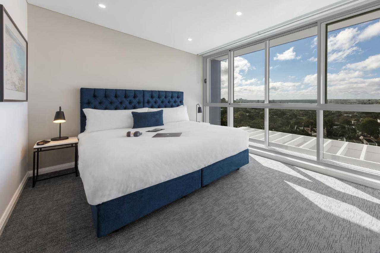 فندق أجنحة ميريتون بوندي جنكشن سيدني المظهر الخارجي الصورة The photo shows a modern and stylish hotel room. It features a comfortable queen-size bed with a blue tufted headboard. The bed is neatly made with white linens and pillows. Next to the bed, there is a small bedside table with a desk lamp. 

On the f