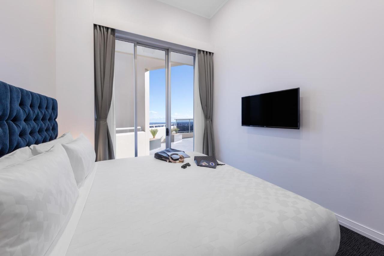 فندق أجنحة ميريتون بوندي جنكشن سيدني المظهر الخارجي الصورة The photo shows a modern hotel room with a large, comfortable bed featuring a blue tufted headboard. There are light-colored walls, and the room is decorated simply. A flat-screen TV is mounted on the wall opposite the bed. The room has large glass d