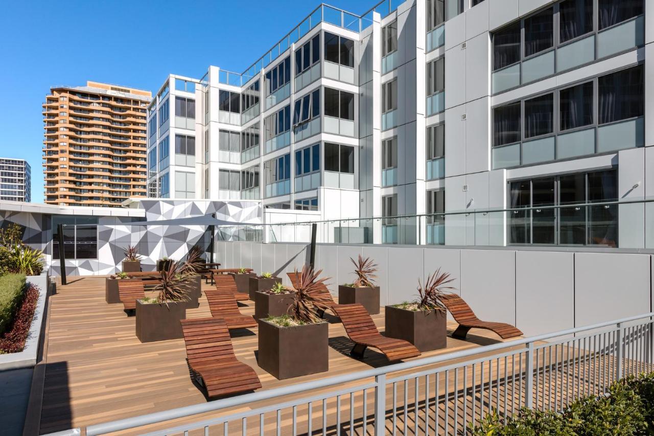 فندق أجنحة ميريتون بوندي جنكشن سيدني المظهر الخارجي الصورة The photo shows a modern outdoor space featuring a wooden deck. There are several lounge chairs arranged around large planter boxes, which contain plants. In the background, there are contemporary residential buildings with large windows, and a geome
