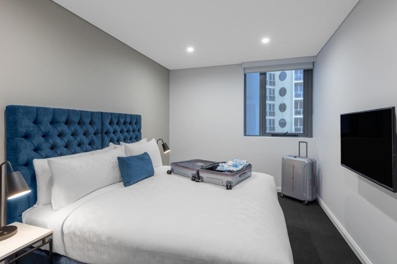 فندق أجنحة ميريتون بوندي جنكشن سيدني المظهر الخارجي الصورة The photo shows a modern hotel room featuring a neatly made bed with a blue tufted headboard. There is a small blue decorative pillow on the bed, and the bedding is white. Next to the bed, there is a nightstand with a lamp. A suitcase is partially op