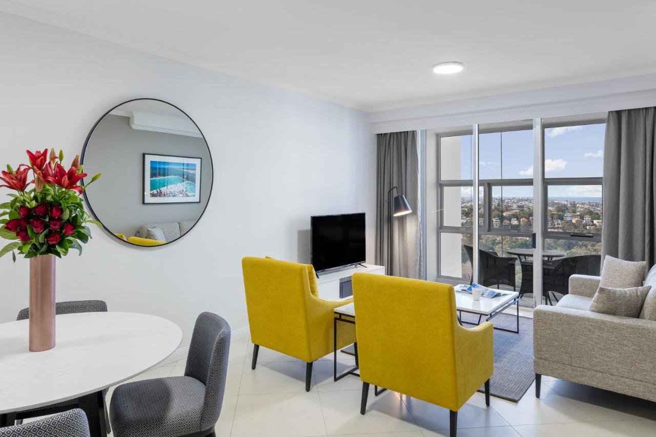 فندق أجنحة ميريتون بوندي جنكشن سيدني المظهر الخارجي الصورة The photo shows a modern living space with a stylish design. There are two bright yellow chairs facing a small glass table, and a comfortable beige sofa to the right. A TV is positioned on the wall in front of the chairs. To the left, there's a dinin