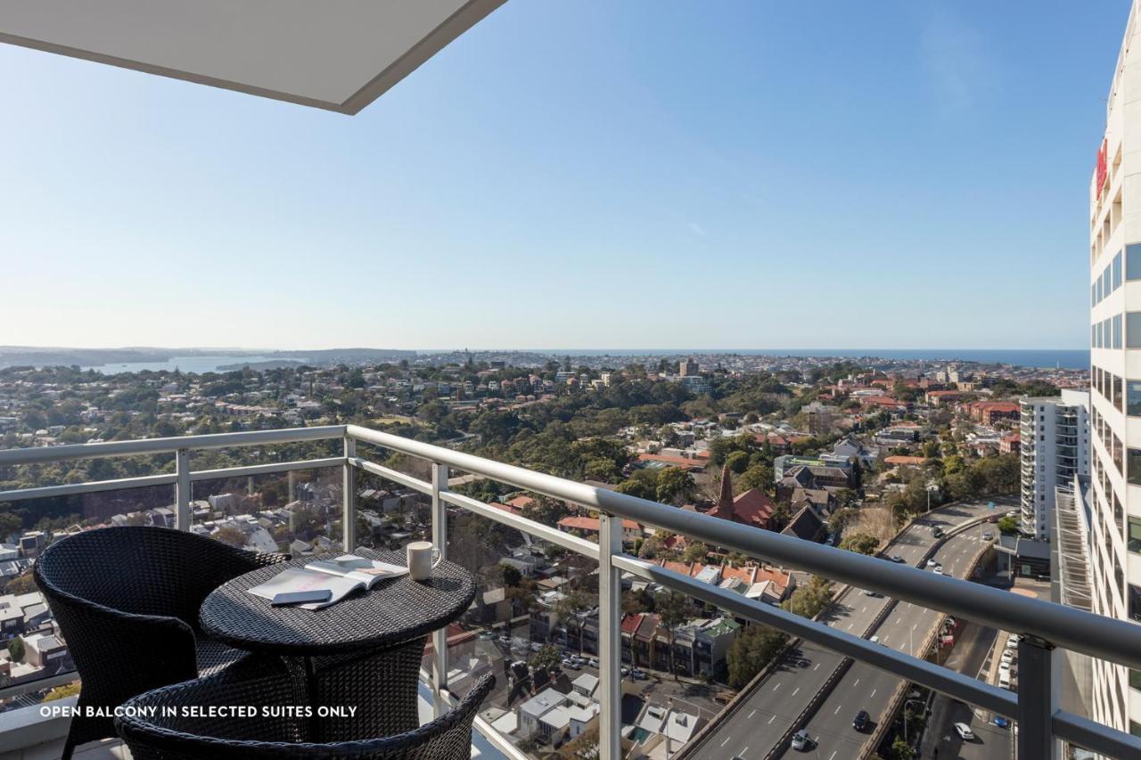 فندق أجنحة ميريتون بوندي جنكشن سيدني المظهر الخارجي الصورة The photo shows a balcony with a view over a cityscape that includes buildings and greenery, extending towards the horizon where the sea is visible. The balcony features a small round table and two black chairs, suggesting a space for relaxation or d