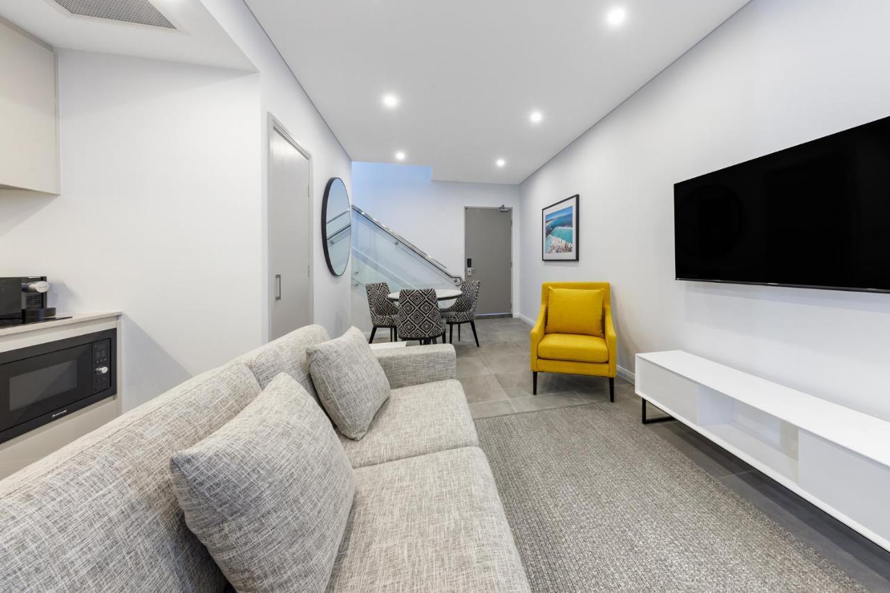 فندق أجنحة ميريتون بوندي جنكشن سيدني المظهر الخارجي الصورة The photo shows a modern and stylish living space. It features a light-colored sofa with cushions, positioned to face a wall-mounted flat-screen TV. There's a yellow accent chair nearby that adds a pop of color to the room. The décor is minimalistic,