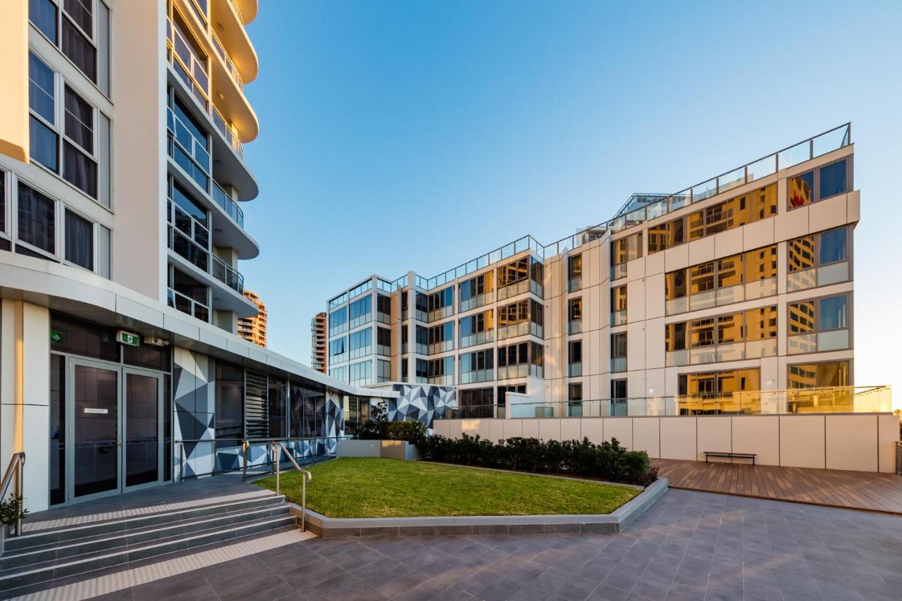 فندق أجنحة ميريتون بوندي جنكشن سيدني المظهر الخارجي الصورة The photo showcases a modern residential area featuring contemporary buildings. In the foreground, there is a landscaped green space surrounded by well-designed architecture. The buildings have large glass windows, reflecting the light, and feature a