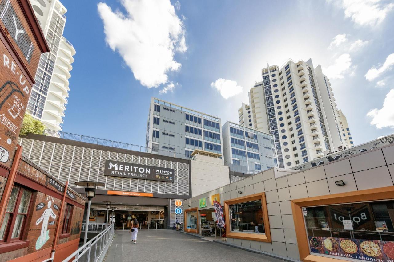 فندق أجنحة ميريتون بوندي جنكشن سيدني المظهر الخارجي الصورة The photo depicts a modern urban environment featuring a building labeled "Meriton Retail Precinct." In the foreground, there are shops with colorful signage, while in the background, tall residential buildings tower over the area. The sky above is b