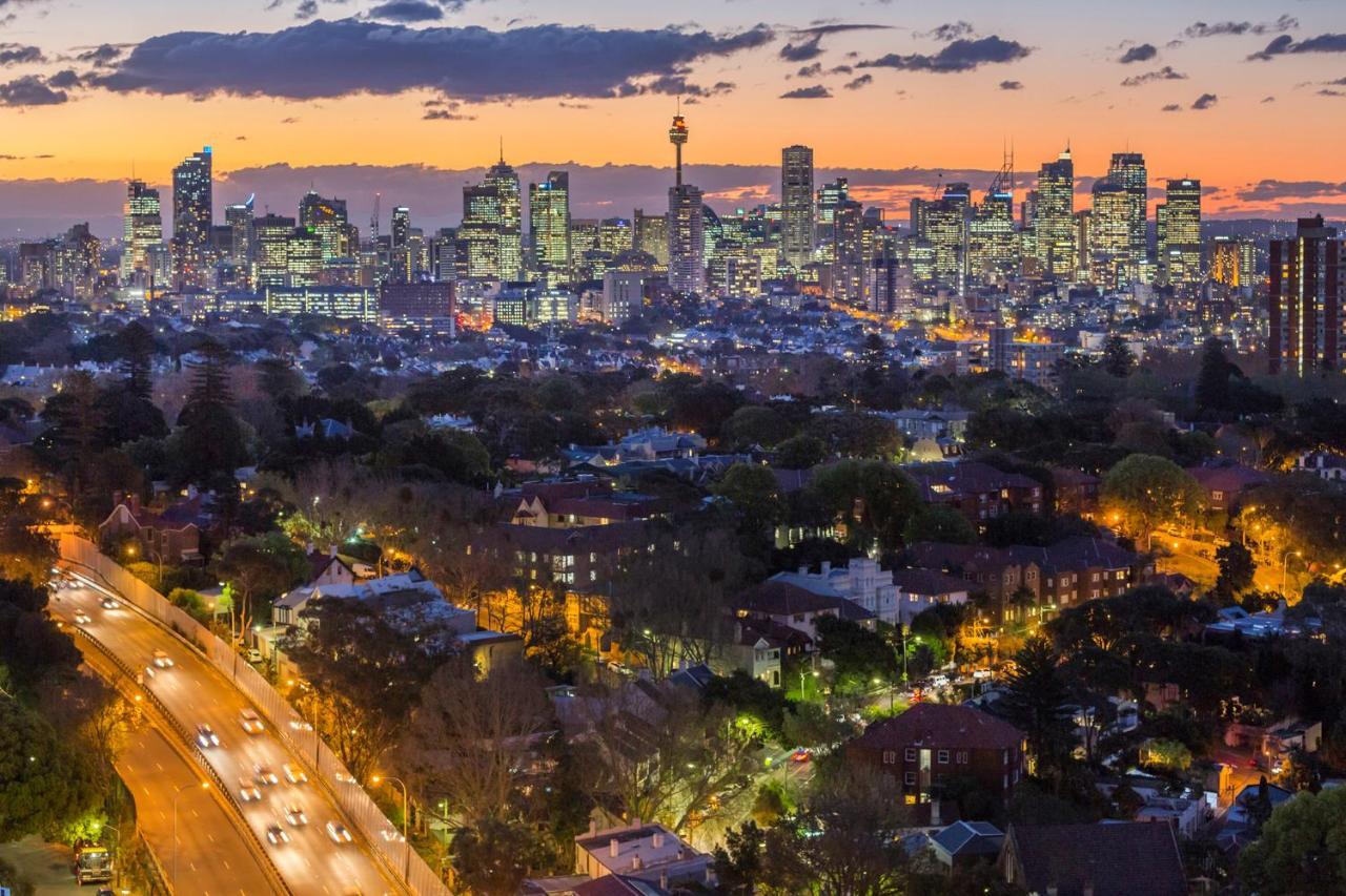 فندق أجنحة ميريتون بوندي جنكشن سيدني المظهر الخارجي الصورة The photo captures a stunning city skyline at dusk, featuring towering skyscrapers adorned with lights against a colorful sunset backdrop. In the foreground, a green area with trees and houses is visible, while a road with cars can be seen winding th