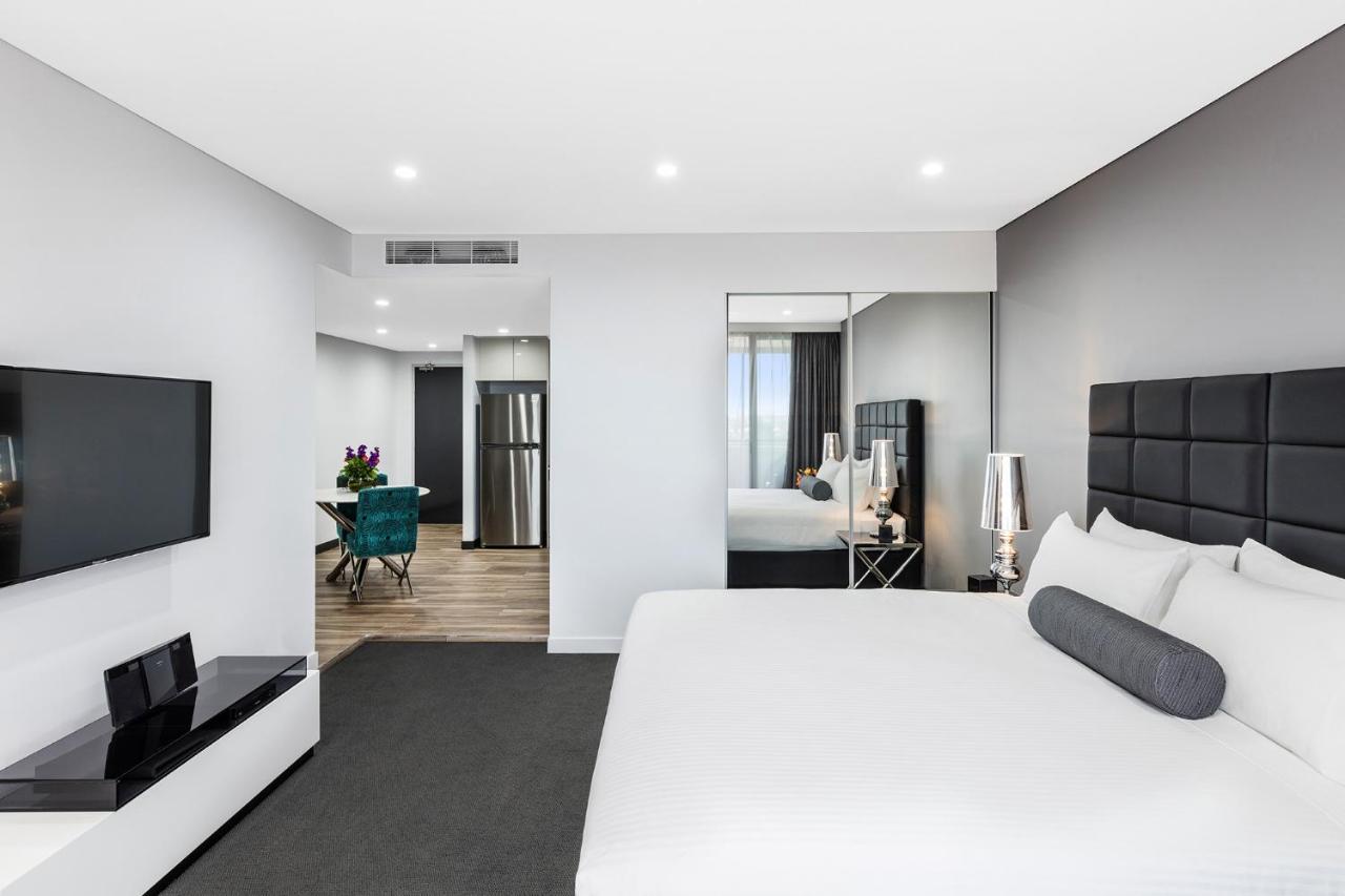 فندق أجنحة ميريتون بوندي جنكشن سيدني المظهر الخارجي الصورة The photo shows a modern hotel room. It features a large bed with a white covering and gray accent pillows. To the left, there's a flat-screen TV mounted on the wall above a sleek white cabinet. The flooring is a light wood tone, and a large mirror i