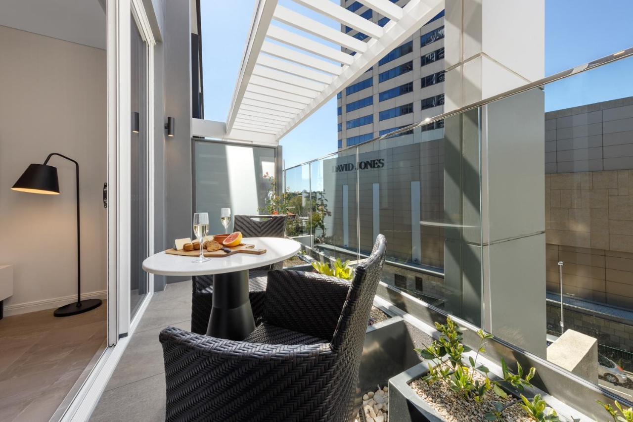 فندق أجنحة ميريتون بوندي جنكشن سيدني المظهر الخارجي الصورة The photo shows a modern balcony area with glass railings. It features a small round table set for two, with a plate of food and drinks. There are two stylish wicker chairs positioned around the table. The space is bright and airy, with sunlight stre