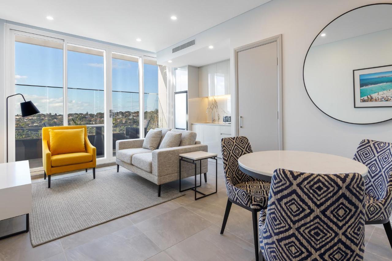 فندق أجنحة ميريتون بوندي جنكشن سيدني المظهر الخارجي الصورة The photo shows a modern and stylish living area. It features a comfortable sofa in a neutral color, accompanied by a bright yellow armchair that adds a pop of color. There's a round dining table with patterned chairs nearby. Large windows allow plen