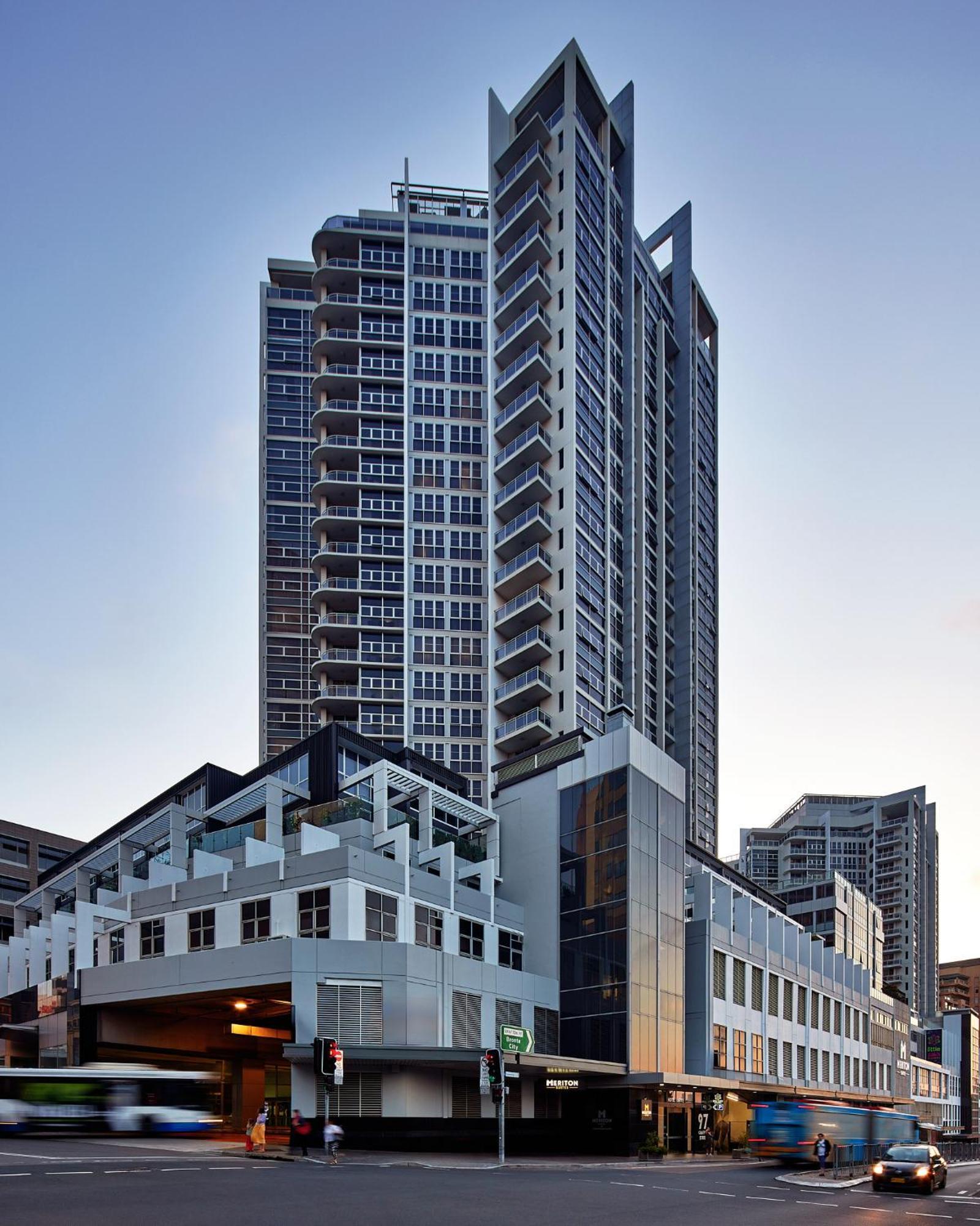 فندق أجنحة ميريتون بوندي جنكشن سيدني المظهر الخارجي الصورة The image shows a modern high-rise building with a sleek design. The structure features a mix of glass and concrete elements, with multiple levels showcasing balconies. The building appears to be part of an urban environment, likely containing reside