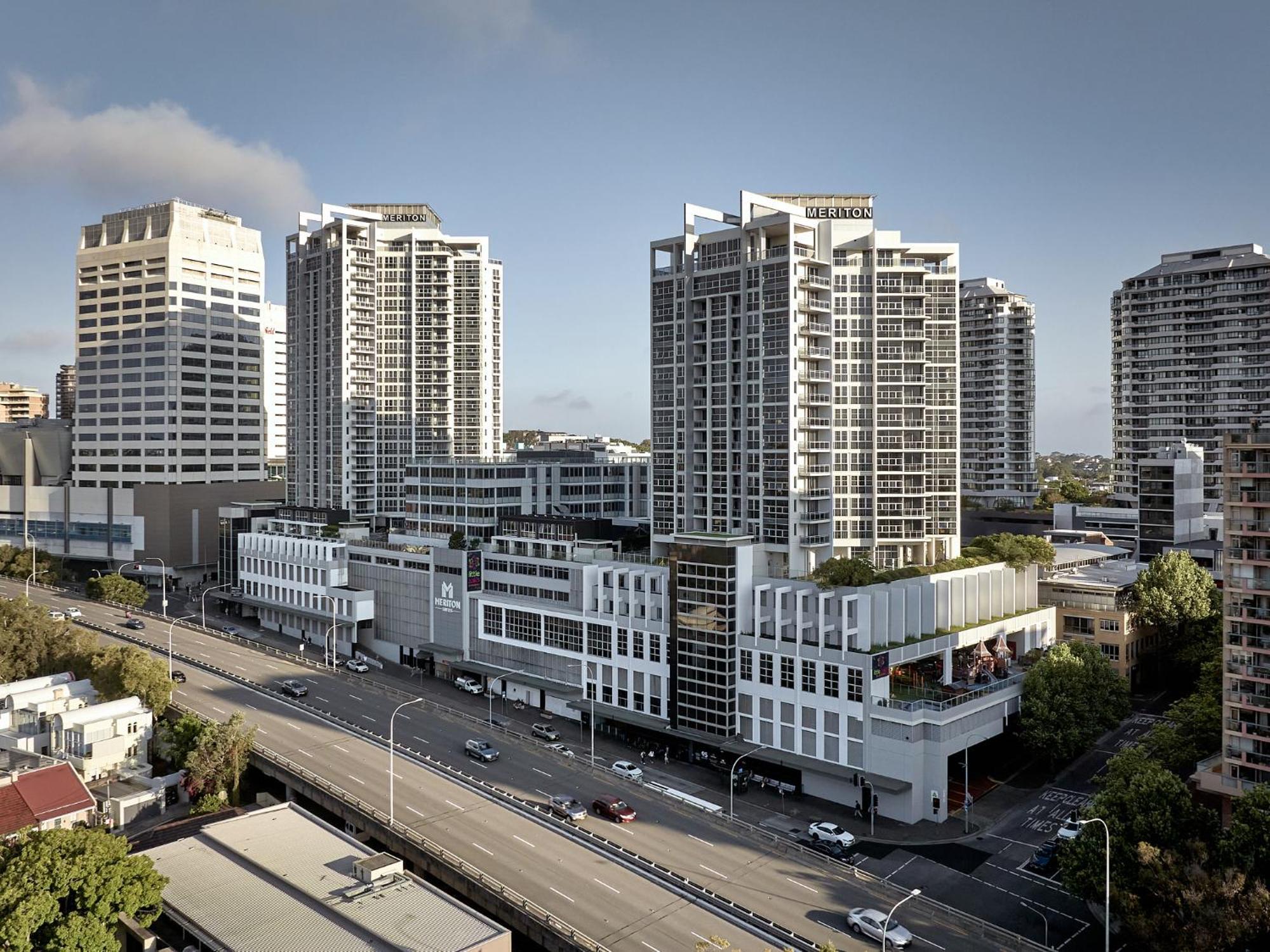 فندق أجنحة ميريتون بوندي جنكشن سيدني المظهر الخارجي الصورة The photo shows a cityscape featuring several tall modern buildings, likely apartments or office complexes. In the foreground, there is a road lined with cars, and the architecture of the buildings varies, with some having sleek glass facades and oth