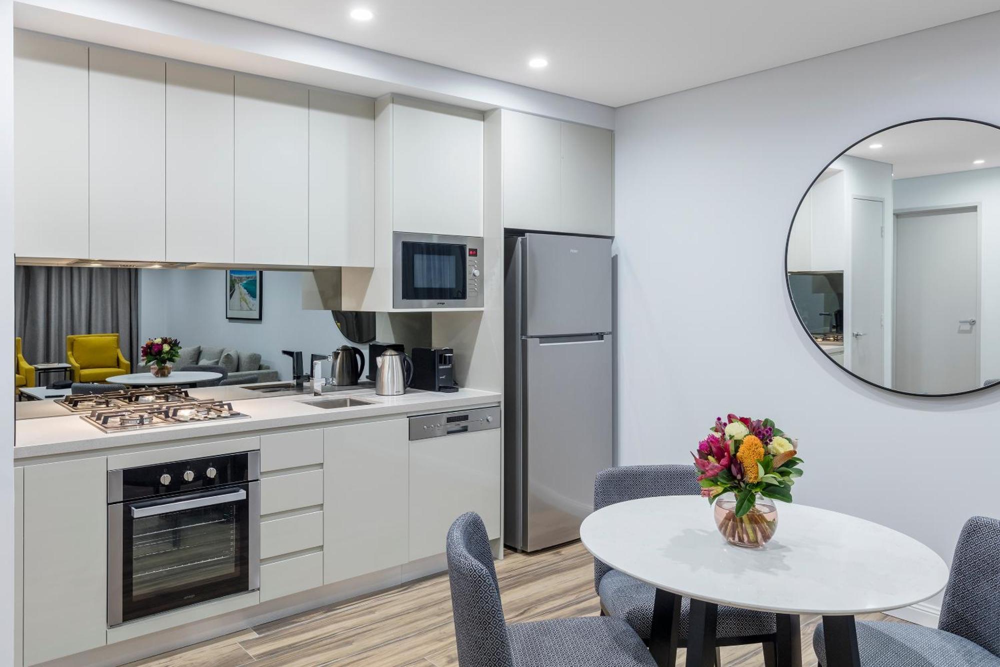 فندق أجنحة ميريتون بوندي جنكشن سيدني المظهر الخارجي الصورة The photo shows a modern kitchen and dining area. The kitchen features light-colored cabinetry with a built-in oven and stovetop, a microwave, and a silver refrigerator. There is a kettle and a coffee maker on the countertop. In the dining area, ther