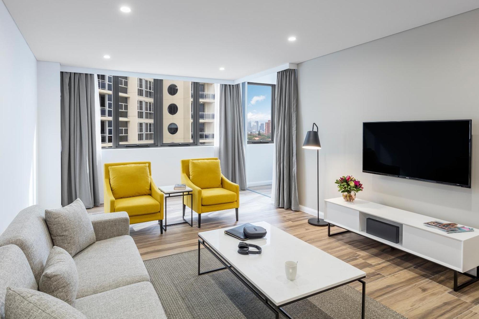 فندق أجنحة ميريتون بوندي جنكشن سيدني المظهر الخارجي الصورة The photo shows a modern, well-lit living room. It features a light gray sofa with a textured fabric. There are two bright yellow armchairs positioned near a window, which allows natural light to enter the space. The room has light gray curtains and 